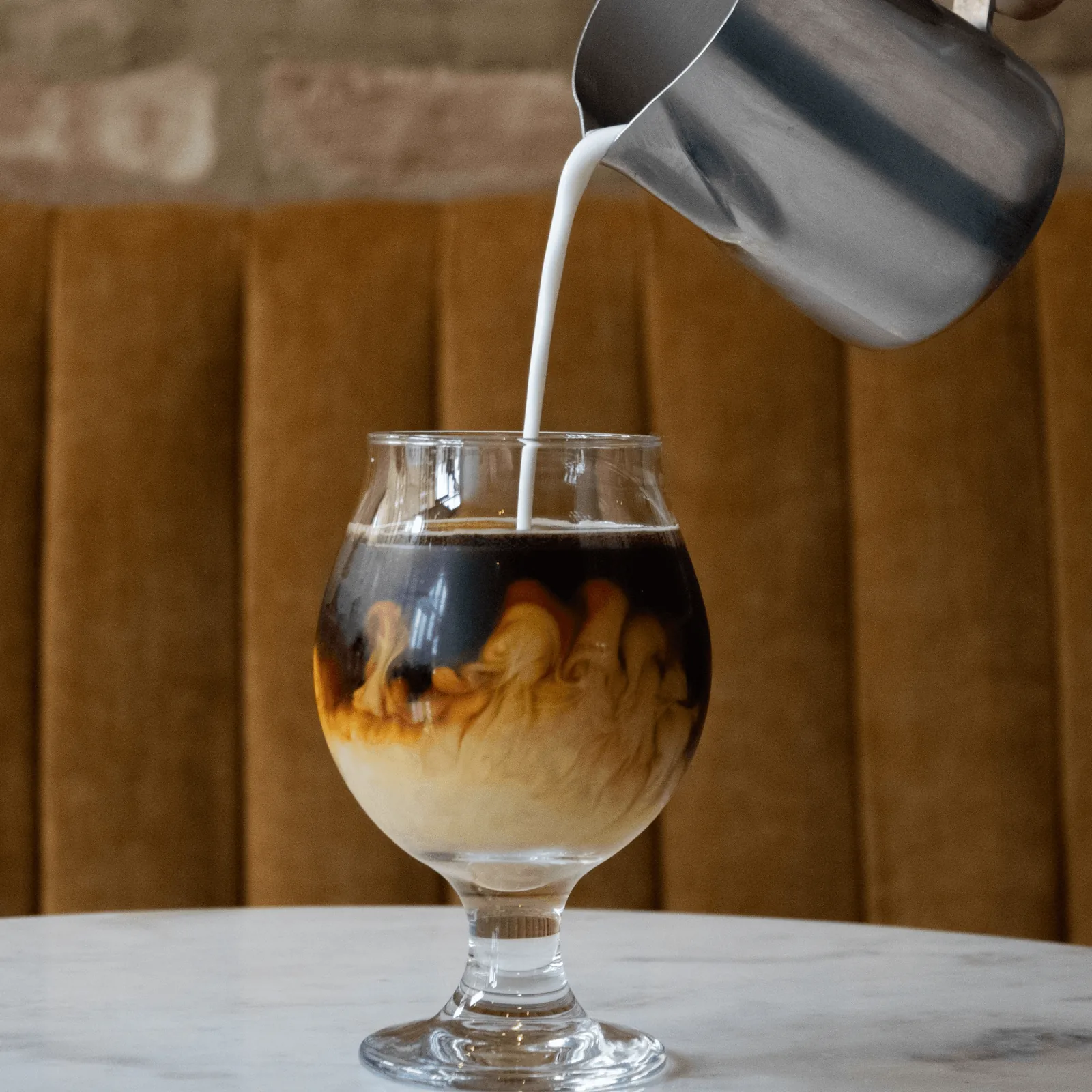 Milk pouring from a metal pitcher into a glass of dark coffee, creating swirls.
