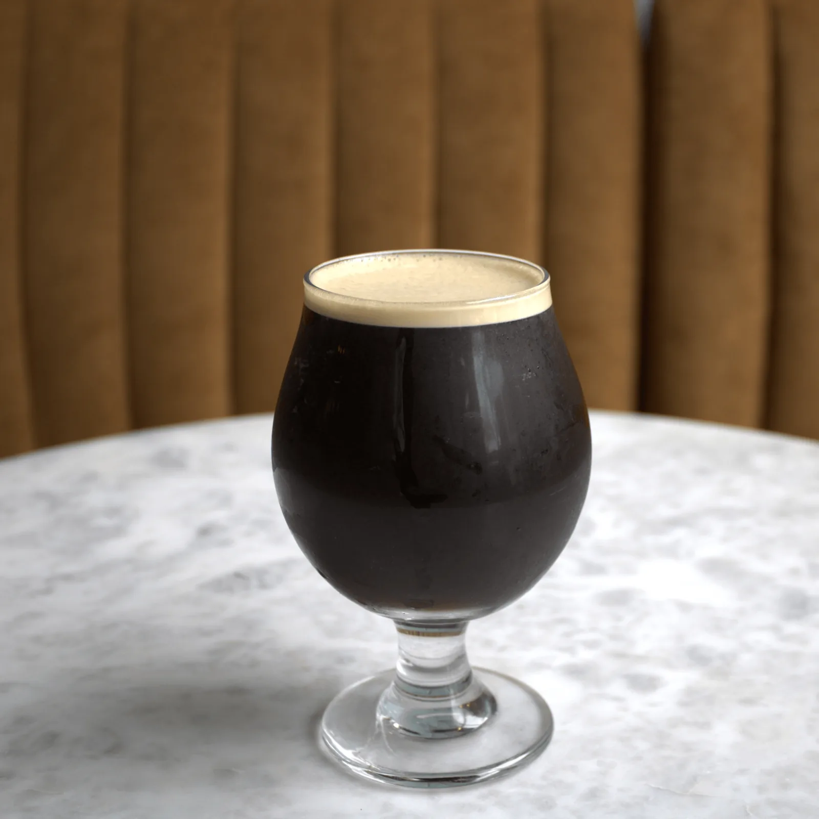A dark coffee drink with a creamy foam top in a stemmed glass on a white marble table.