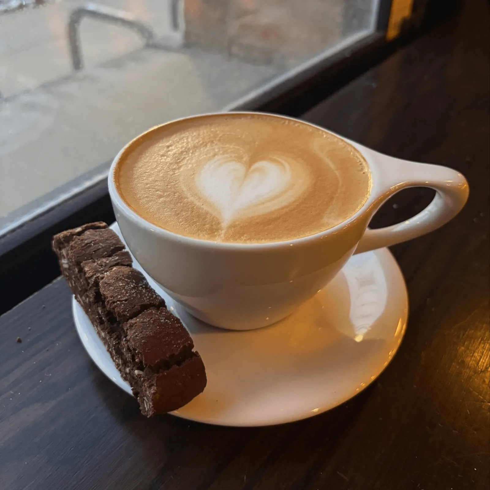 Ipsento 606 latte with heart art and a chocolate biscotti on a saucer by a window.
