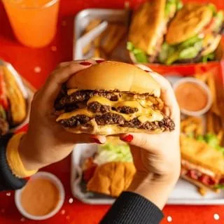 Hands holding a double cheeseburger with melted cheese and caramelized onions, surrounded by other burgers and fries.