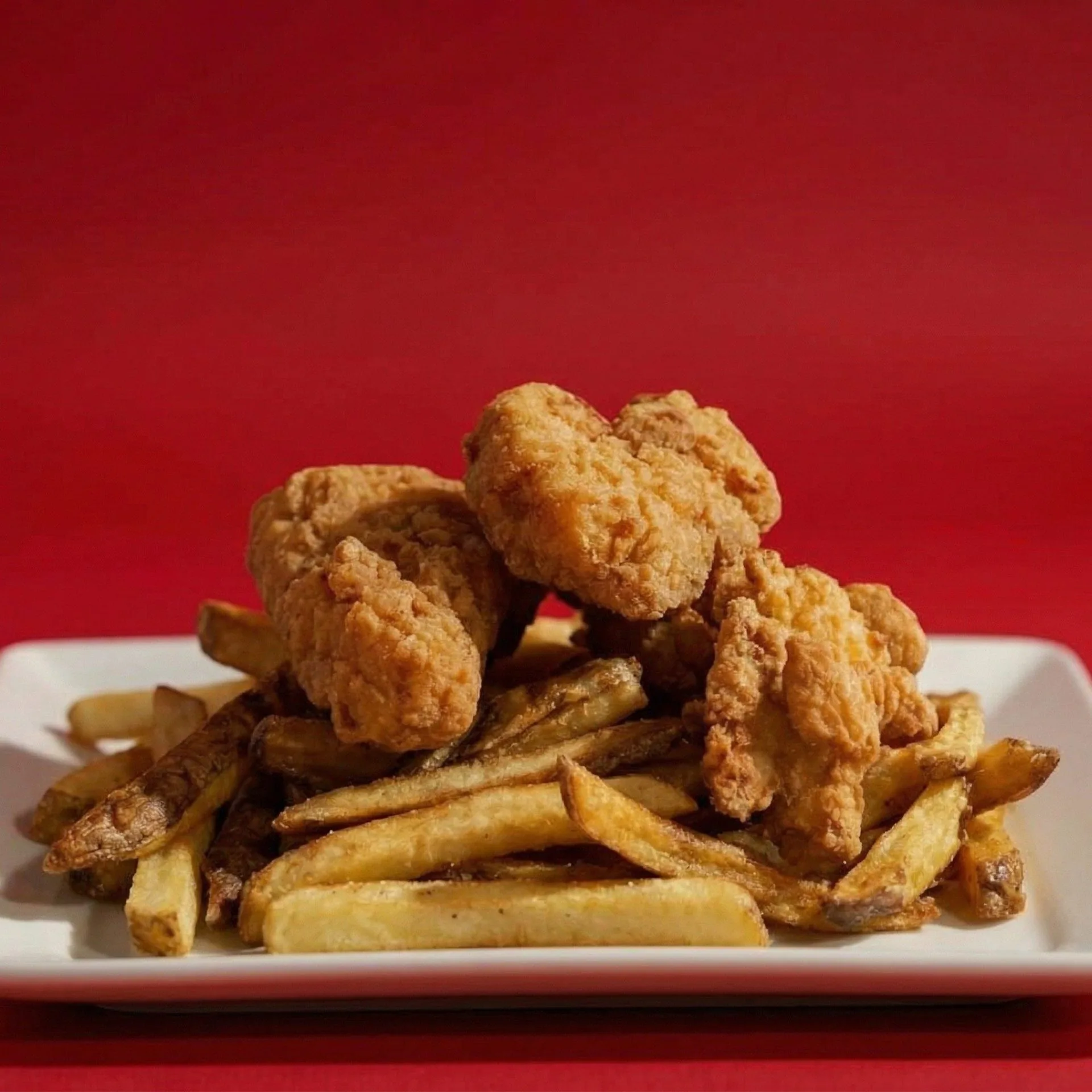 A white plate piled high with golden-brown fried chicken tenders and crispy french fries against a red background.
