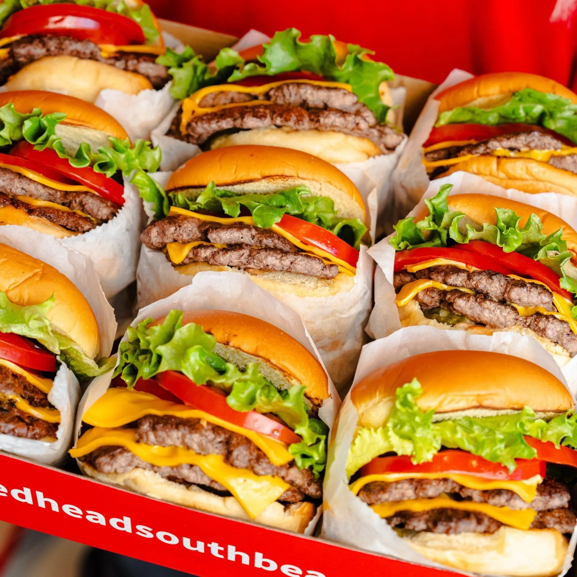 A box filled with many double cheeseburgers, each wrapped in white paper with lettuce and tomato.