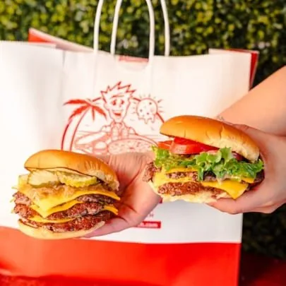 Two hands hold two RedHead Burger double cheeseburgers, one with pickles and onions, the other with lettuce and tomato.